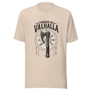 May include: Beige t-shirt featuring a black graphic of a Viking axe and the words "FORGED IN VALHALLA". The axe is centered over a circular design with runes. The shirt has short sleeves and a crew neck.
