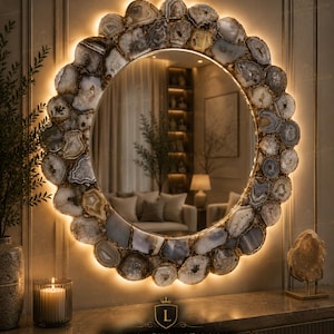 Puede incluir: Un espejo redondo con un marco decorativo de piedras de ágata natural en tonos grises, blancos y marrones. El marco está iluminado con una luz cálida y suave. El espejo forma parte de una colección de decoración del hogar de lujo.