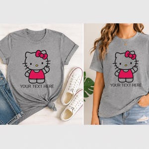Pode incluir: Duas camisetas cinzentas com um gato de desenho animado com la&ccedil;o e vestido rosa. O texto "YOUR TEXT HERE" est&aacute; abaixo do gato. Uma camisa &eacute; estilizada com jeans e t&ecirc;nis. A outra &eacute; usada por uma pessoa.