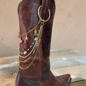 May include: A dark brown leather cowboy boot adorned with a multi-layered gold-tone chain accessory. The accessory features a large circular ring, a small gold cowboy boot charm, a starfish charm, a pink heart charm, white beads, and rhinestones.