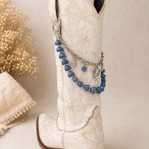 May include: A cream-colored western boot featuring intricate tonal stitching and a decorative boot chain. The accessory includes a strand of blue heart-shaped beads, a silver-toned metal chain, a small horseshoe charm, a star, and a bull skull charm.