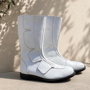 Puede incluir: Un par de botas de cuero blanco con suelas negras. Las botas tienen una altura media de pantorrilla y presentan costuras decorativas y un cierre de velcro. Las botas están sobre un fondo neutro con sombras.