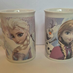 May include: Two white ceramic mugs featuring characters from the movie Frozen. The left mug displays Elsa with a blonde braid, while the right mug features Anna and Olaf. Both mugs have a curved handle and a smooth, glossy finish with light blue accents.