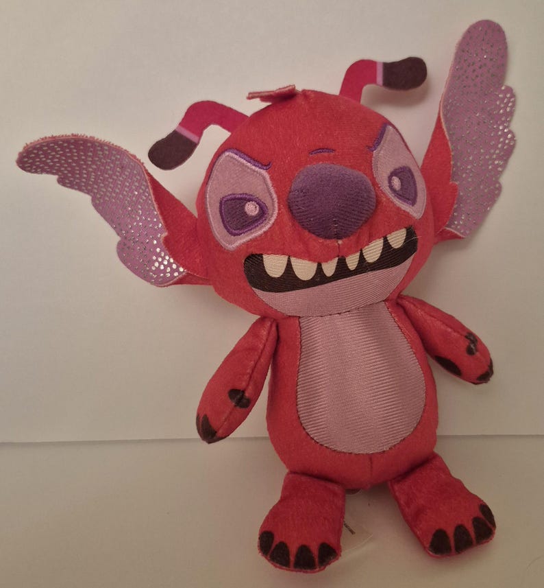 May include: A red plush toy character with large, expressive eyes, a purple nose, and a wide grin revealing white teeth. The character has large, glittery purple wings and is posed with its arms and legs extended. The toy is a popular character from a children's animated movie.