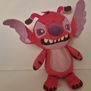 May include: A red plush toy character with large, expressive eyes, a purple nose, and a wide grin revealing white teeth. The character has large ears and wings with a glittery, iridescent finish. The toy is a soft, stuffed animal.