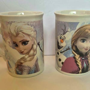 May include: Two white ceramic mugs featuring characters from the movie Frozen. The left mug displays Elsa with her blonde braided hair, while the right mug features Anna and Olaf the snowman against a light blue and white winter-themed background.