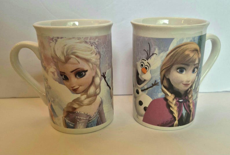May include: Two white ceramic mugs featuring characters from the movie Frozen. The left mug displays Elsa with her blonde braided hair, while the right mug features Anna and Olaf the snowman against a light blue and white winter-themed background.