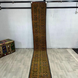 May include: A long, narrow runner rug featuring a repeating geometric pattern in shades of dark orange, olive green, and deep blue. The rug has a distinct border and a central vertical panel with intricate, symmetrical motifs throughout its length.