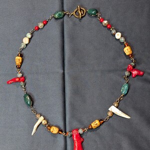 May include: A necklace featuring a gold-toned toggle clasp, interspersed with red coral-like branches, yellow skull-shaped beads, smooth green stones, white tusk-shaped charms, and small brown and clear beads connected by gold-toned metal links.