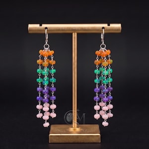 May include: A pair of dangle earrings featuring multiple strands of small, round, multi-colored beads in shades of orange, green, purple, and light pink. The beads are strung on thin silver-toned metal chains, creating a layered, cascading effect.