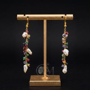 May include: A pair of gold-toned drop earrings featuring a cascading arrangement of small, multicoloured gemstones and white baroque pearls. The earrings are suspended from a gold-toned metal T-bar display stand against a dark background.
