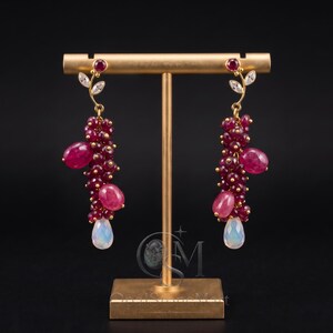 Peut inclure: Une paire de boucles d'oreilles dorées composées de grappes de perles de rubis facettées rouge foncé, de cabochons de rubis lisses plus grands et d'une pierre de lune irisée en forme de goutte à la base, surmontées d'un rubis et deux cristaux.