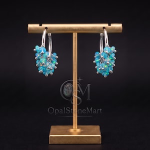 May include: A pair of silver hoop earrings featuring clusters of small, round, iridescent blue opal beads. The earrings are displayed on a gold-toned T-shaped stand. The text OpalStoneMart is visible in the center of the stand, along with a logo.