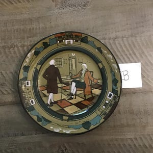 May include: A decorative plate with a painted scene of two figures in a room. The plate has a green border with architectural and tree designs. The central image features a checkered floor and a table.