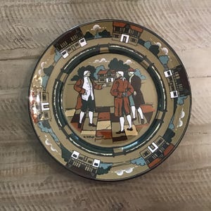 May include: A decorative plate with a historical scene featuring three figures in period clothing. The plate has a tan background with a dark green border and a repeating pattern of buildings and trees. The plate is round.