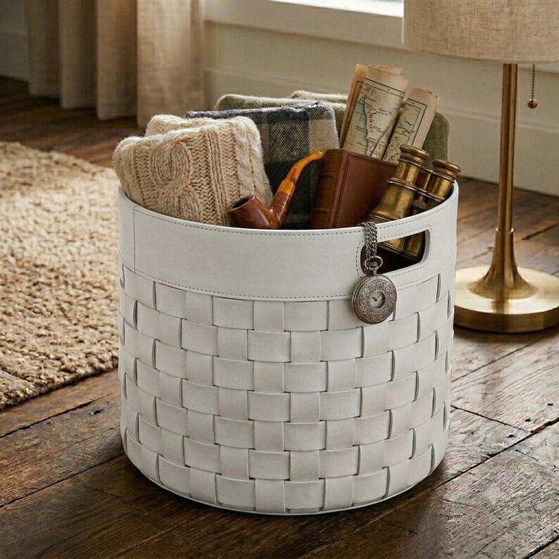 Puede incluir: Una cesta de almacenamiento redonda de tejido blanco con un borde superior liso y un dije de reloj de bolsillo plateado en el lateral. La cesta contiene un tejido beige de punto trenzado, un libro encuadernado en cuero marr&oacute;n, una pipa y binoculares.