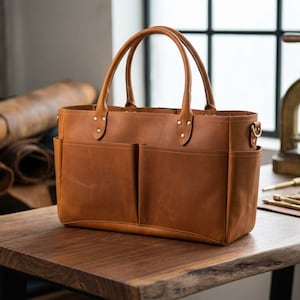 May include: A brown leather tote bag with two front pockets and two top handles. The bag has brass hardware and is sitting on a wooden table. Rolls of leather are in the background.