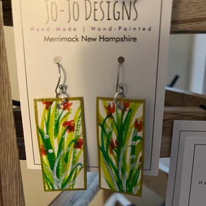 May include: Hand-painted rectangular earrings with a floral design. The earrings feature a white background with green grass, red flowers, and a gold border. The text "Jo-Jo Designs" is visible on the tag.