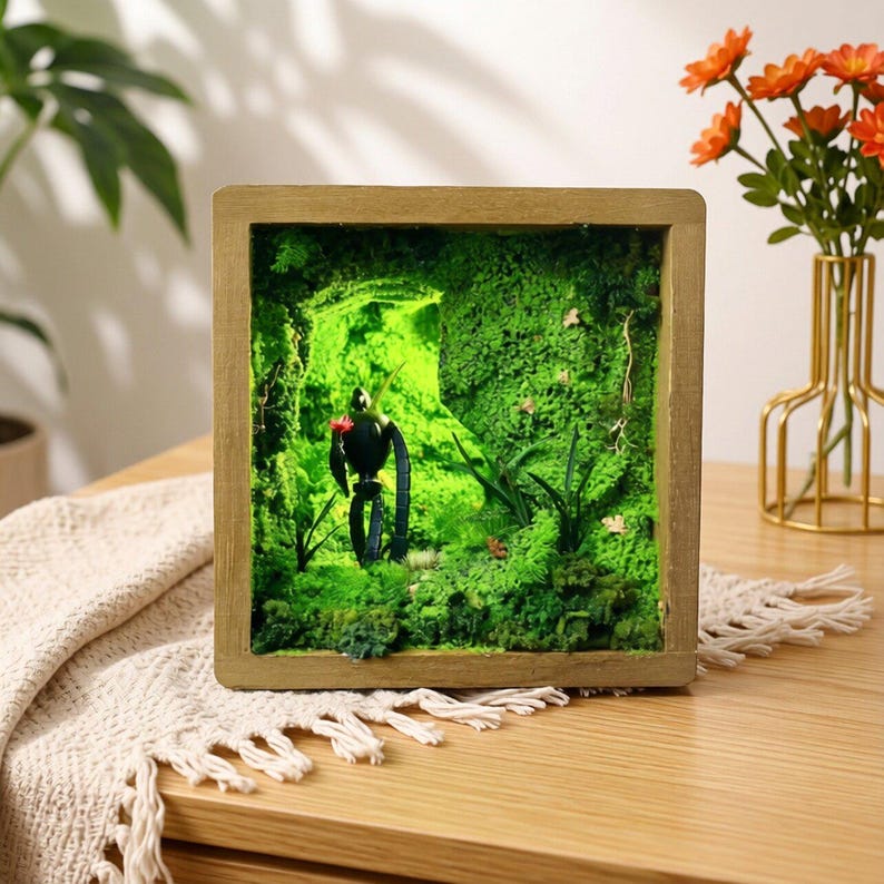 May include: A framed diorama featuring a miniature toucan sculpture in a lush, green moss environment. The toucan holds a red flower. The wooden frame surrounds the miniature forest scene, creating a unique decorative piece.