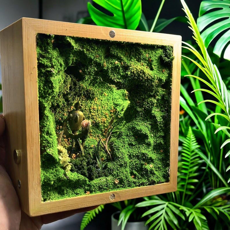 May include: A square wooden box with a miniature moss garden inside. The moss is a vibrant green, and a small, detailed sculpture is nestled within the scene. The box has a natural wood finish, and the overall aesthetic is reminiscent of a terrarium.