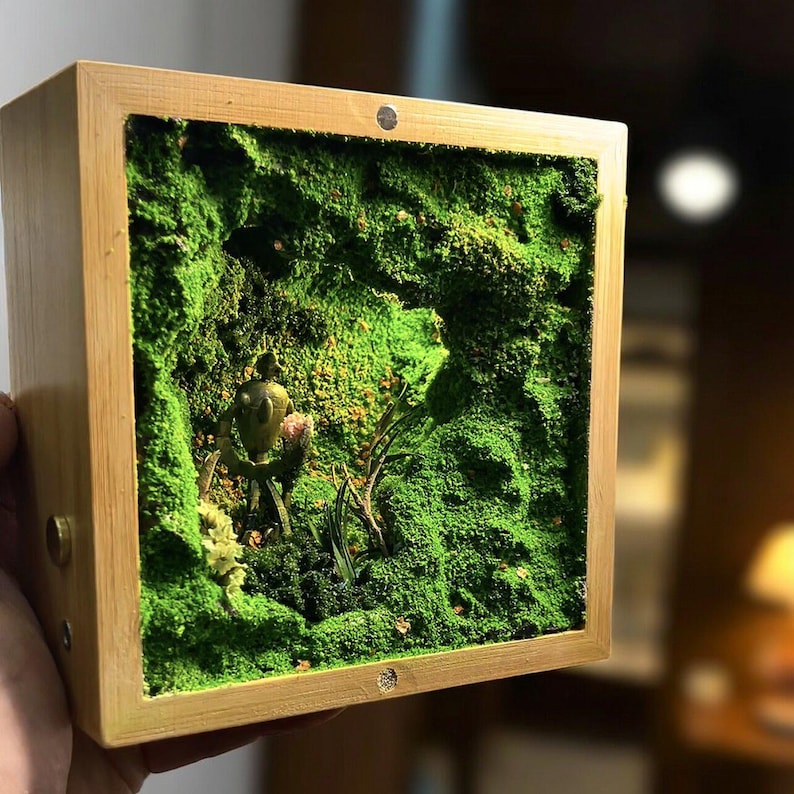 May include: A small, square wooden box with a miniature moss garden inside. The box has a natural wood finish and a small metal accent. The moss is a vibrant green, and a small figurine is visible within the miniature landscape.