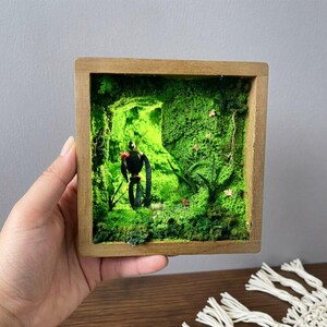 May include: A miniature diorama in a wooden frame featuring a black robot figure in a vibrant green mossy landscape. The scene includes small plants and a touch of red, creating a whimsical, nature-inspired art piece. The frame is held by a hand.
