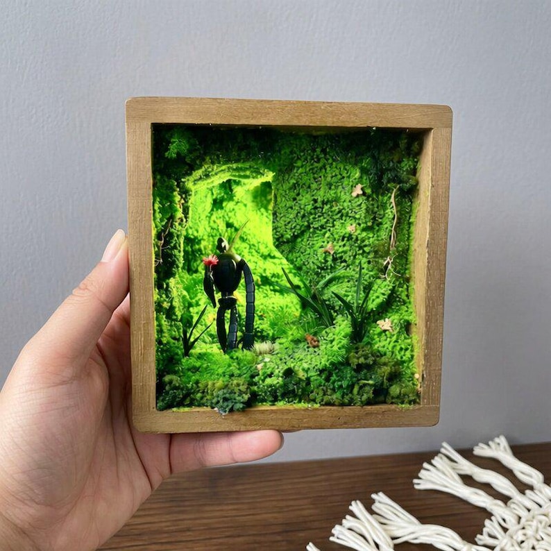 May include: A miniature diorama in a wooden frame featuring a black robot figure in a vibrant green mossy landscape. The scene includes small plants and a touch of red, creating a whimsical, nature-inspired art piece. The frame is held by a hand.