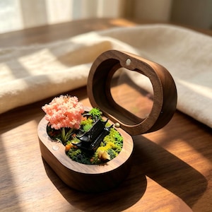 May include: An oval wooden box with a hinged lid containing a miniature diorama. The scene features a small black pagoda structure nestled among green moss, tiny colorful foliage, and a delicate pink flowering tree, all encased in wood.