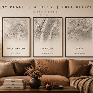 May include: Three framed map prints of Ho Chi Minh City, New York, and Osaka, displayed above a brown sofa. The maps are in black and white, with city names and coordinates below. The text "ANY PLACE | 3 FOR 2 | FREE DELIVERY" is above the prints.