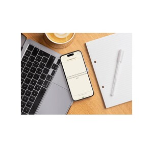 May include: A laptop, smartphone, notepad, pen, and a cup of coffee are arranged on a wooden surface. The smartphone displays text from Revelation 21:4. The scene suggests a workspace or study environment.