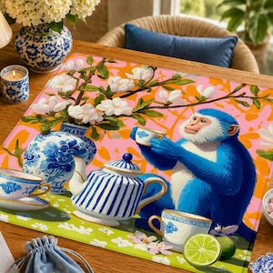 May include: A rectangular placemat featuring a vibrant illustration of a blue monkey holding a teacup. The scene includes a blue and white striped teapot, a matching teacup, a blue and white floral vase with white flowers, and a sliced lime.