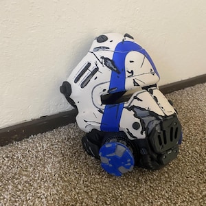 May include: A white and blue helmet featuring a weathered, battle-worn finish with black scuff marks and scratches. The helmet includes a prominent blue stripe across the top, a black faceplate section, and circular side components with blue accents.