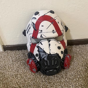 May include: A white full-face helmet featuring a bold red arch design across the forehead and nose area. The surface displays black distressed weathering marks and scratches. It includes a black respirator-style mouthpiece and red circular side vents.