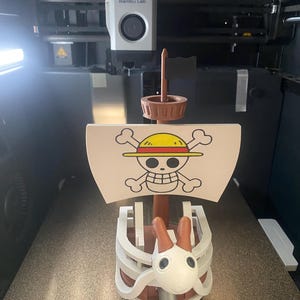 May include: A 3D-printed model of a ship from the anime series One Piece. The ship features a white sail with the Jolly Roger symbol, a brown mast, and a white and brown figurehead. The model is on a 3D printer.