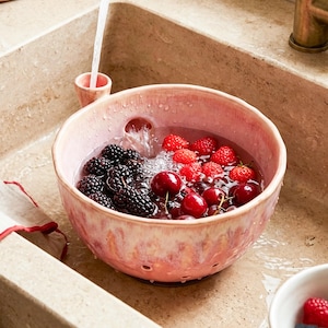 May include: A pink ceramic colander bowl filled with fresh blackberries, raspberries, and cherries being rinsed under a stream of water from a brass kitchen faucet. The bowl features a small integrated funnel for water intake and drainage holes.