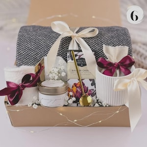 May include: A gift basket containing a grey and white blanket, a candle, chocolate, and a small white container, all tied with burgundy bows. The basket is decorated with white flowers and a gold spoon, tied with a cream ribbon.