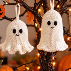 May include: Two off-white knitted ghost ornaments with black yarn eyes and a small stitched smile. Each ghost has a loop at the top for hanging and a flared, wavy bottom edge. The texture is consistent with hand-knitted yarn, creating a soft appearance.