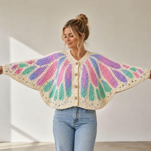 Puede incluir: Un cárdigan de ganchillo color crema con una silueta ancha en forma de alas y un patrón vibrante de pétalos radiantes en tonos rosa pastel, lavanda y verde menta. La prenda tiene un cuello en V y tres botones de color marrón claro.