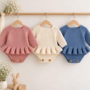 May include: Three long-sleeve crochet rompers hanging on wooden hangers. Each romper features a ruffled waistline, a round neckline, and two small wooden buttons at the bottom closure. The rompers are available in dusty pink, cream, and dusty blue colors.