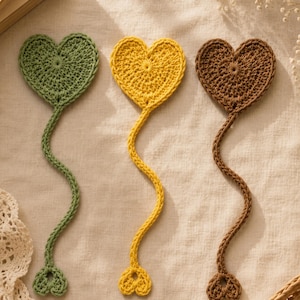 May include: Three crocheted bookmarks in sage green, mustard yellow, and brown. Each bookmark features a large heart-shaped top connected by a long, thin, braided cord to a smaller, matching heart-shaped charm at the bottom end of the cord.