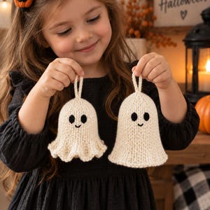 May include: Two off-white knitted ghost ornaments featuring black embroidered eyes and a small smile. Each ghost has a small loop at the top for hanging. The texture of the yarn is visible, showing a simple knit stitch pattern on both figures.