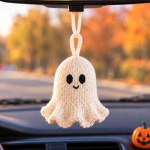 May include: A cream-colored, hand-knitted ghost ornament hanging from a rearview mirror. The ghost features a rounded head, a flared base, and two black embroidered oval eyes with a small, simple black embroidered smile centered on its face.