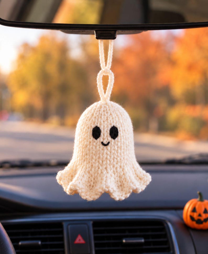 May include: A cream-colored, hand-knitted ghost ornament hanging from a rearview mirror. The ghost features a rounded head, a flared base, and two black embroidered oval eyes with a small, simple black embroidered smile centered on its face.
