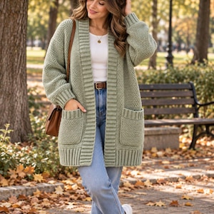May include: A sage green open-front cardigan featuring a chunky knit texture, long sleeves with ribbed cuffs, two large patch pockets, and a wide ribbed placket. The garment is worn over a white top, blue jeans with a brown belt, and white sneakers.
