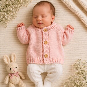 May include: A pale pink knitted long-sleeve cardigan featuring three round wooden buttons down the centre front, paired with white knitted footed leggings. A small cream-coloured crocheted rabbit toy with a pink bow sits beside the clothing.