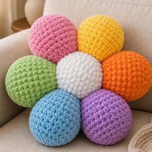 Könnte beinhalten: Eine Anordnung von acht bunten, runden, gehäkelten Kissen in einem blütenartigen Muster. Die Kissen sind in den Farbtönen Rosa, Gelb, Orange, Grün, Weiß, Blau und Lila gehalten. Die Kissen liegen auf einem beigen Sofa.