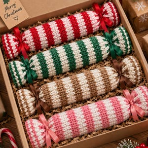 May include: Four knitted candy-shaped ornaments in a cardboard box. Each ornament features horizontal stripes in red, green, brown, or pink, alternating with white. Each end is tied with a matching satin ribbon. A tag reads Merry Christmas.