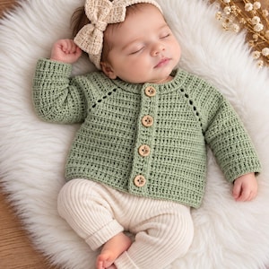 Puede incluir: Un cárdigan de manga larga en color verde salvia con un patrón de punto texturizado y cuatro botones redondos de madera en la parte delantera. La prenda se combina con leggings acanalados de color crema y una diadema de ganchillo a juego con lazo.