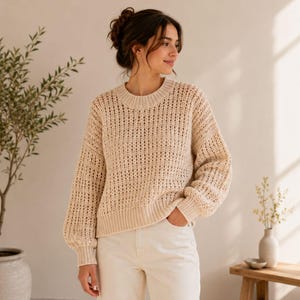 May include: A beige long-sleeved jumper featuring an open-knit, grid-like texture with a ribbed crew neckline, cuffs, and hem, paired with off-white denim trousers. The jumper has a relaxed fit with dropped shoulders and a consistent light colour.