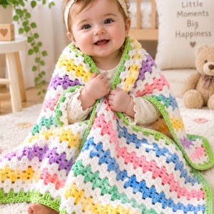 May include: A baby wearing a floral headband and a cream-colored long-sleeved shirt, wrapped in a handmade crochet blanket. The blanket features a colorful granny square pattern with stripes of white, yellow, pink, blue, purple, and light green.